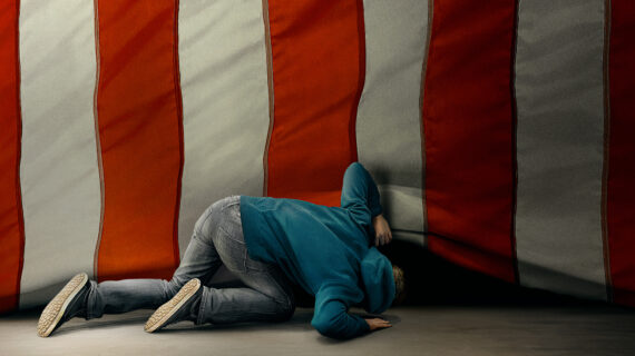 A person in a blue hoodie and gray jeans kneels on the floor, peering underneath the edge of a large American flag.