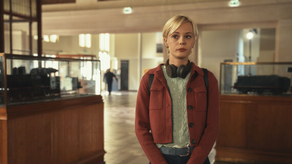 A young woman wearing headphones around her neck stands in a softly lit museum gallery with glass display cases and wooden exhibits in the background.