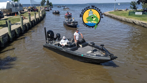 A fishing boat with two men navigates a canal during a sunny day. Several boats are in the background. A "Walleye Weekend" logo is also present.