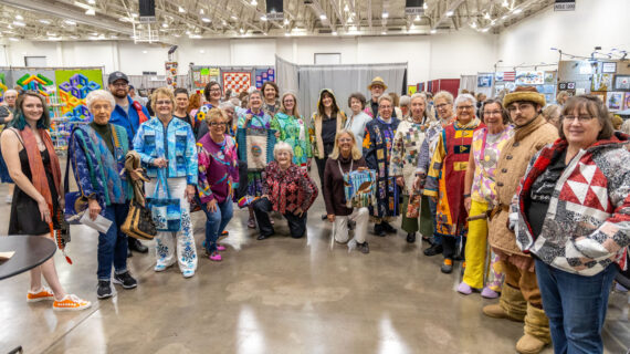 A happy group of people pose with their colorful quilted garments in an expo center.