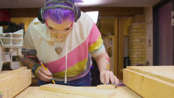 A tattooed woodworker with purple hair, safety glasses, and ear protection marks a measurement on a piece of lumber in a workshop.