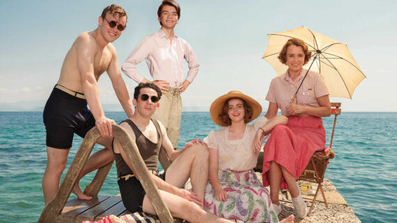 Five people in vintage 1930s-style swimwear and summer clothing pose together on a wooden pier beside a calm blue sea.
