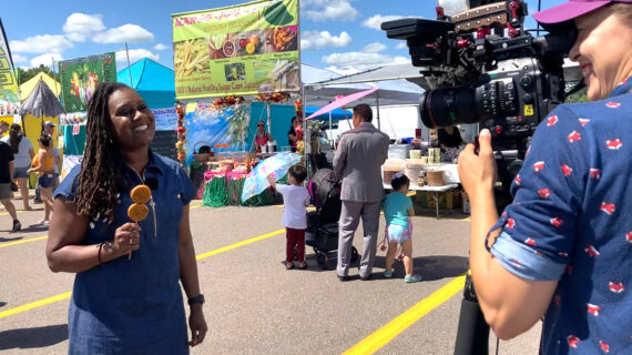 Wisconsin Life host Angela Fitzgerald smiles, holding food on sticks, at an outdoor market at the Northern Wisconsin State Fair with colorful stalls. A camera operator films her, creating a lively, cheerful scene.