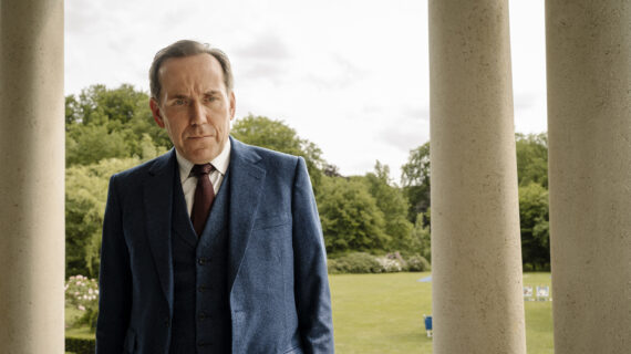 A man in a three-piece suit standing alongside columns of a building outdoors stares off-camera with a serious expression.