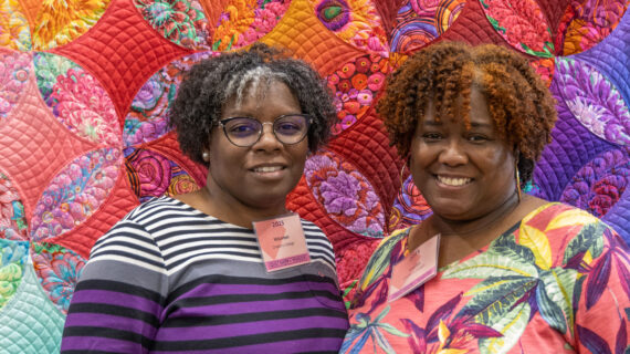 Two women stand smiling in front of a colorful quilt.