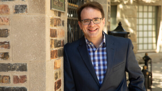 WPR host Jason Heilman shares his favorite PBS Wisconsin classical music programs WPR "Drivetime Classics" host Jason Heinemann smiling in glasses, navy blazer, and blue plaid shirt leans against a brick building, creating a warm, approachable outdoor portrait.