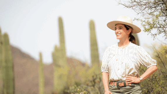 Woman in a blouse and cowboy hat stares into the distance against a backdrop of desert and saguaro cacti.