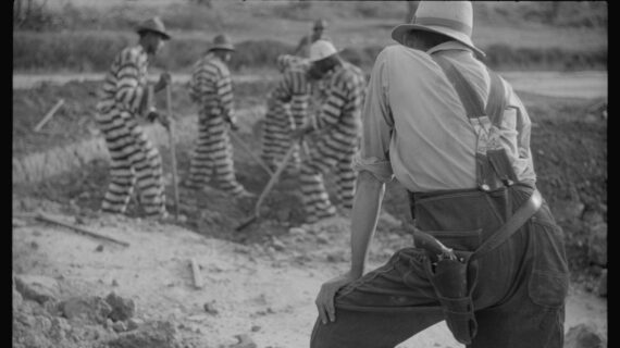 A man with a pistol on his hip oversees a group of prisoners wearing striped one-piece outfits digging a trench.